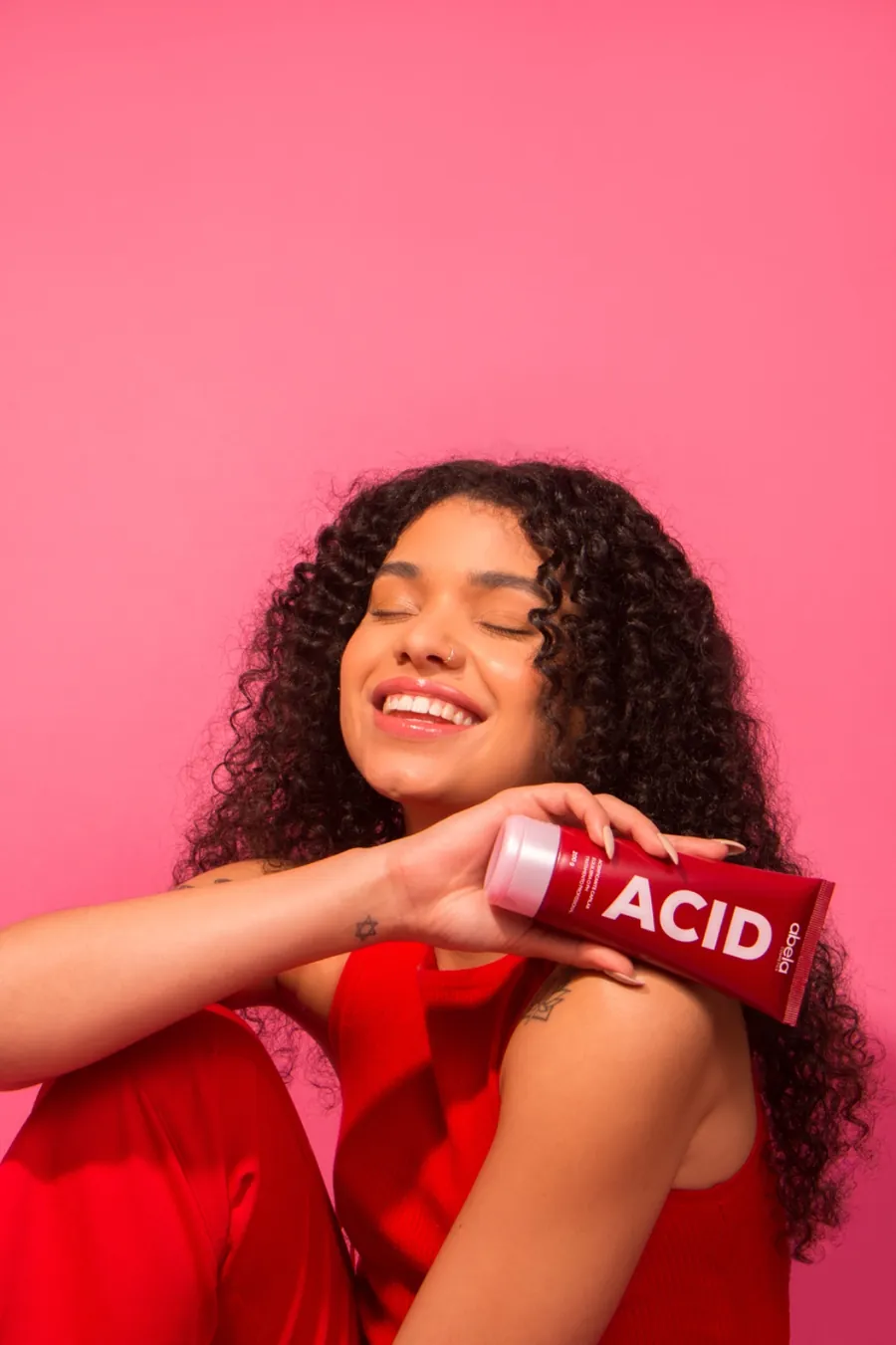 Criadora de conteúdo sorrindo e segurando o hidratante ACID da Abela Cosmetics, em fundo rosa.