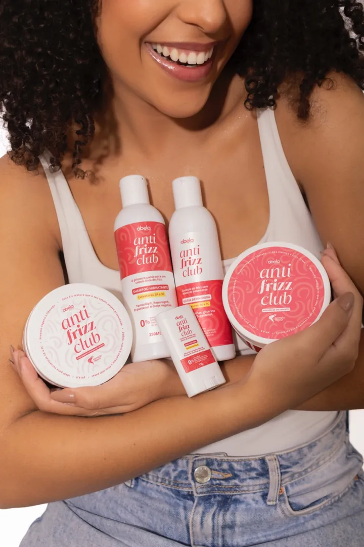 Kit Anti Frizz Club da Abela: shampoo hidratante, geleia definidora, máscara de tratamento e cera, segurados por uma modelo de cabelo cacheado.